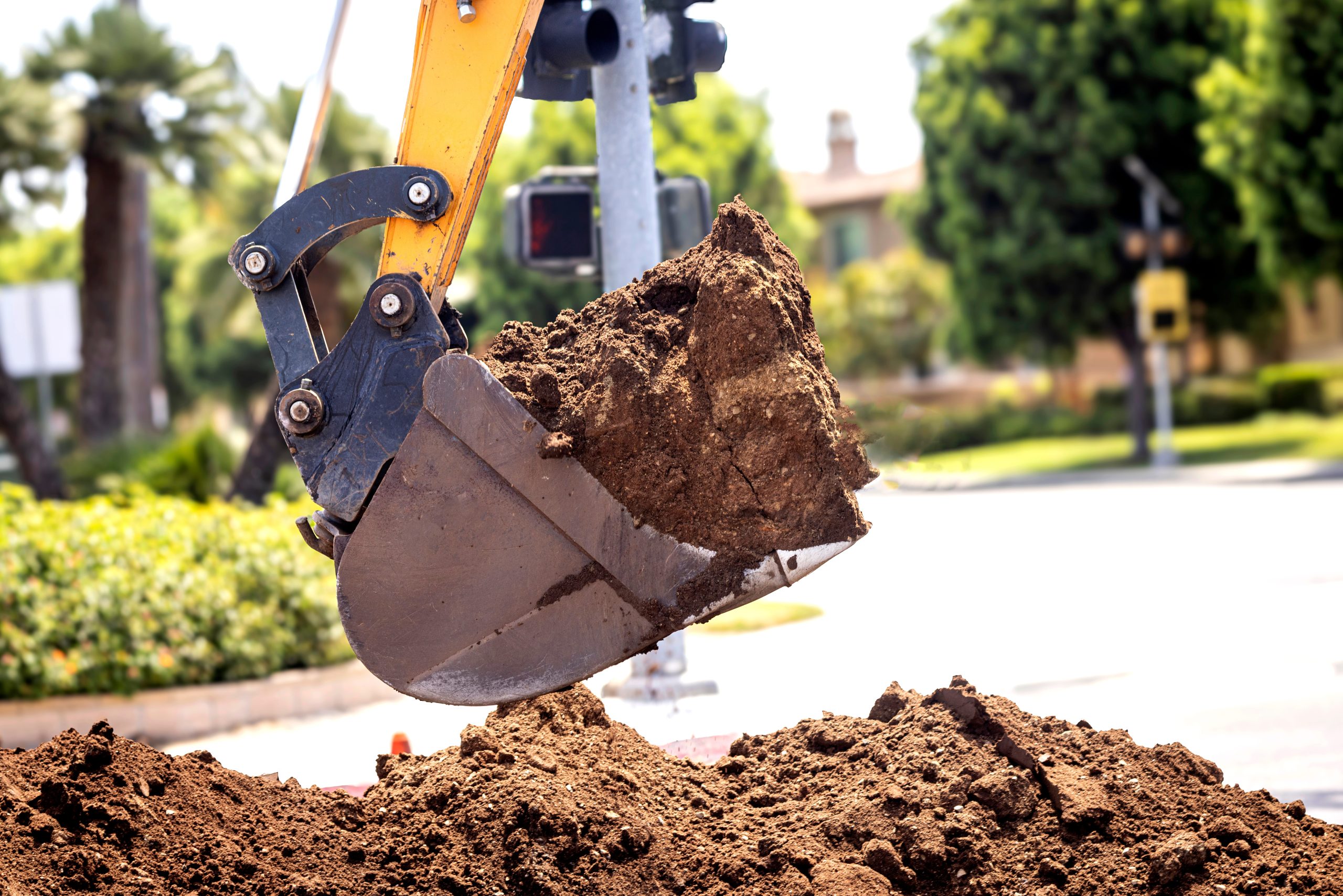 Close up of a backhoe scoop bucket filled with dirt . The work is being done at a residential intersection. Excavator arm lifting dirt near the street with trees and buildings in the background.