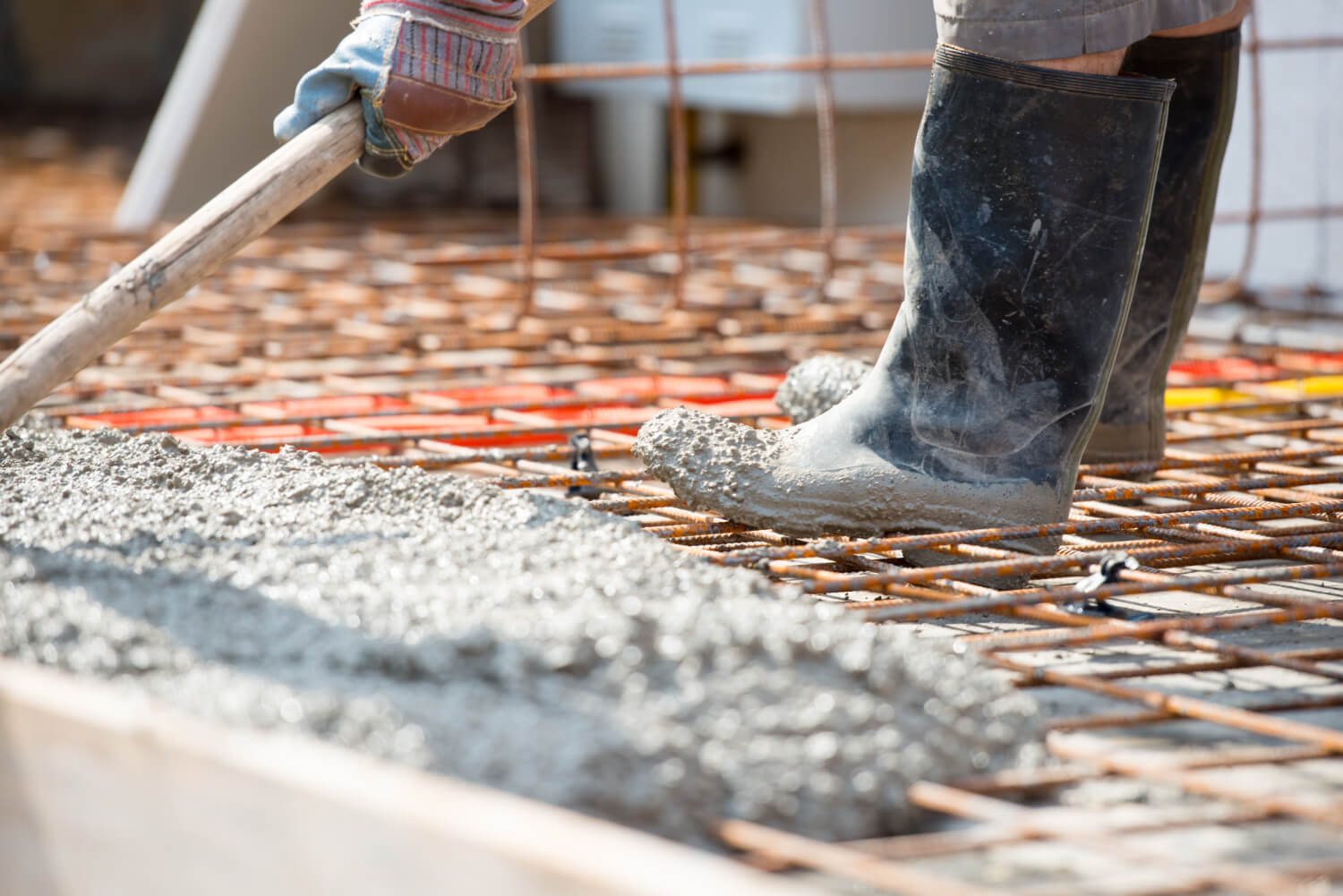 Worker in rubber boots spreading wet concrete over steel rebar at a construction site.