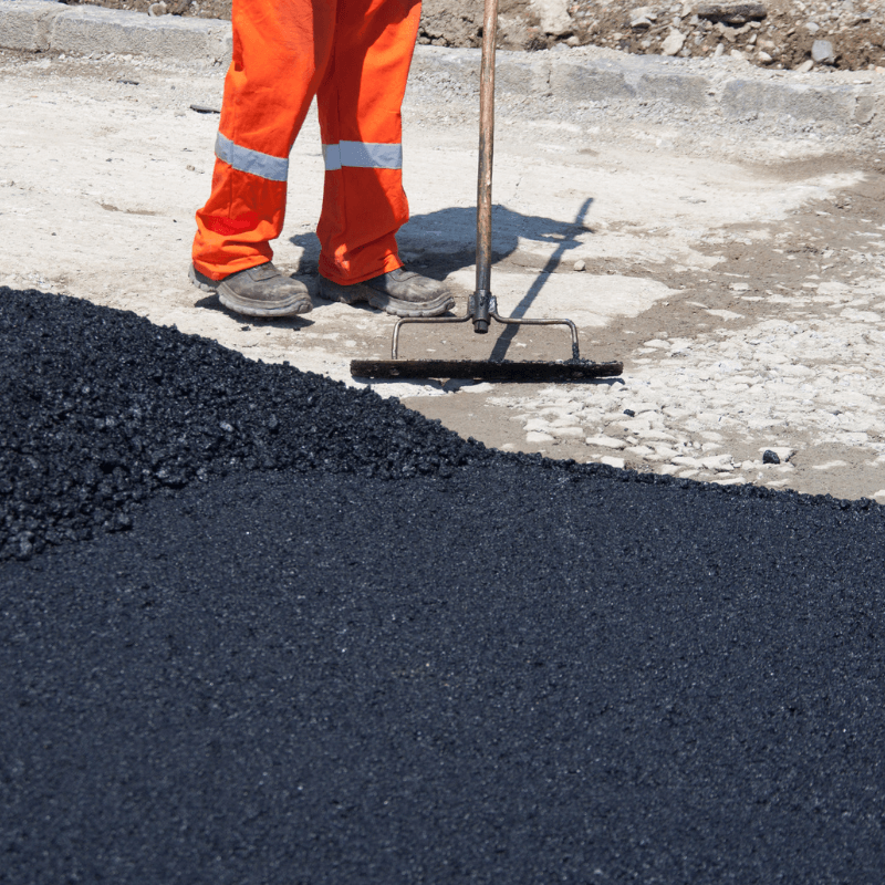 Worker in orange pants levels hot asphalt on a road with a rake during road construction.