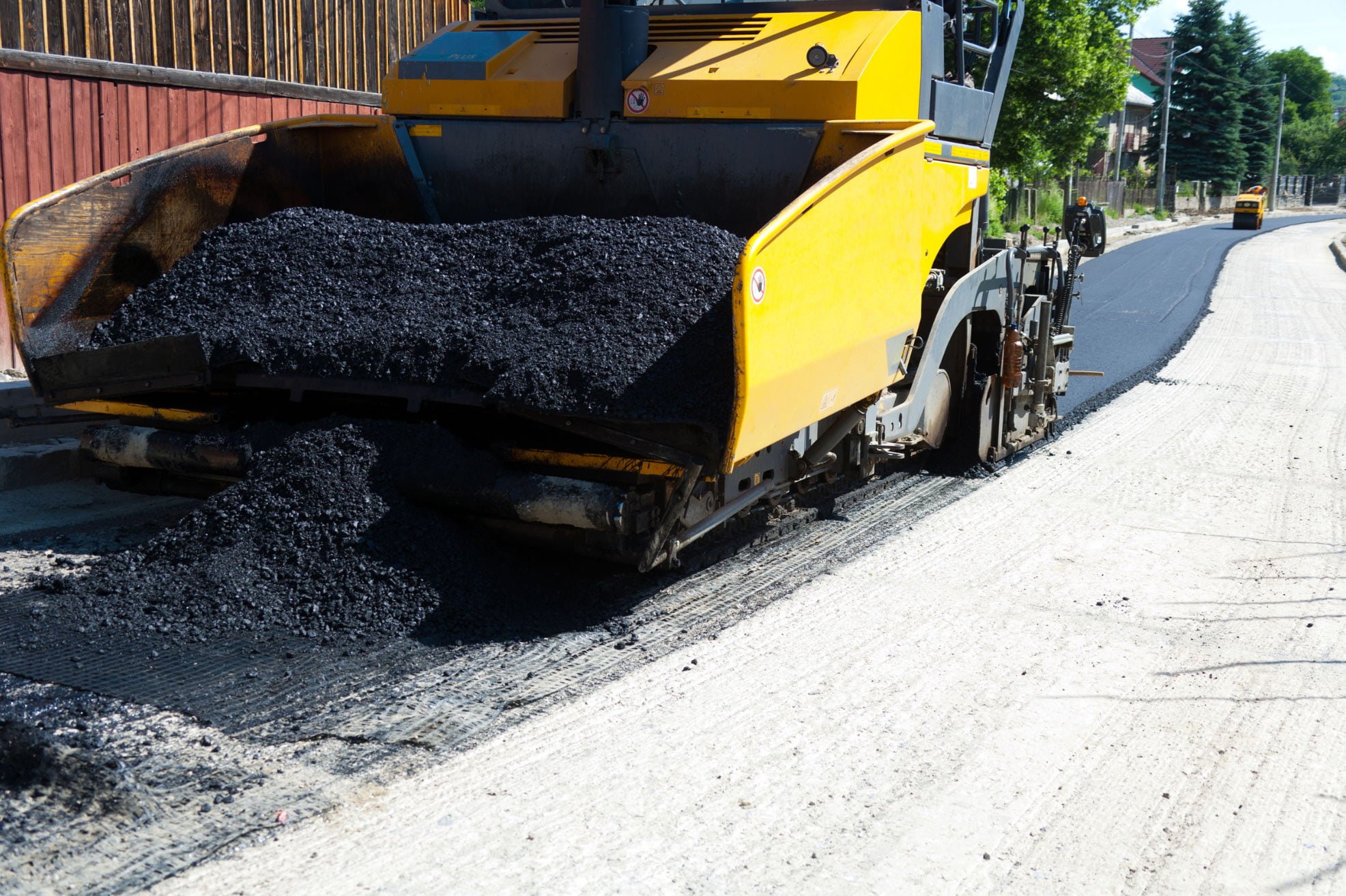 A road paver machine spreads fresh asphalt on a street under construction on a sunny day.