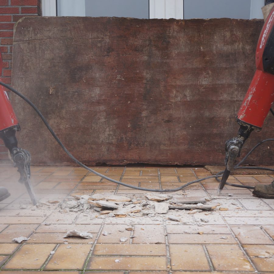 Two workers use jackhammers to break up a tiled surface, with dust and debris scattered around.