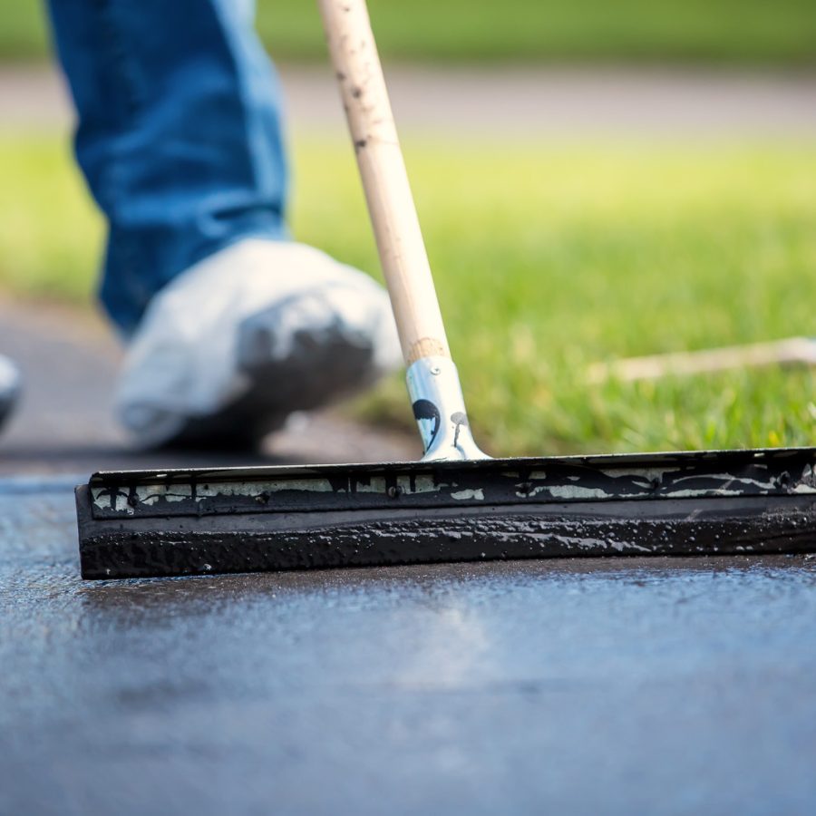 Person applying sealant to a driveway with a squeegee near green grass, wearing blue pants and shoe covers.
