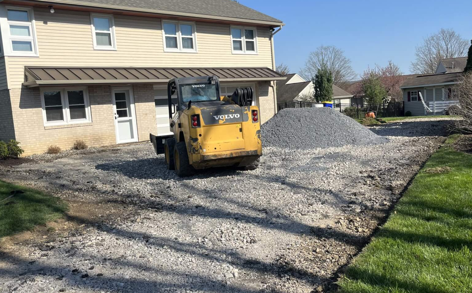 A small Volvo loader on gravel in front of a house, with a pile of stones nearby.