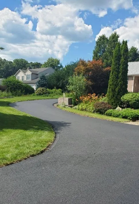 A freshly paved driveway curves past green lawns and shrubs toward houses under a blue sky with clouds.